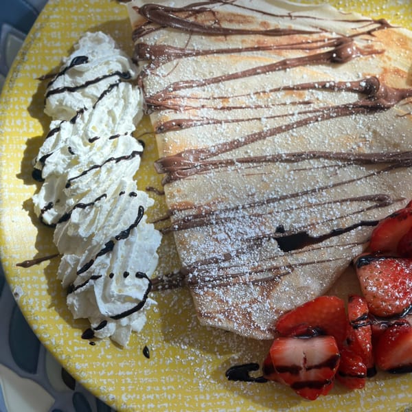 Manasiri Crepe with chocolate drizzle, powdered sugar, whipped cream, and fresh strawberries on a yellow plate.