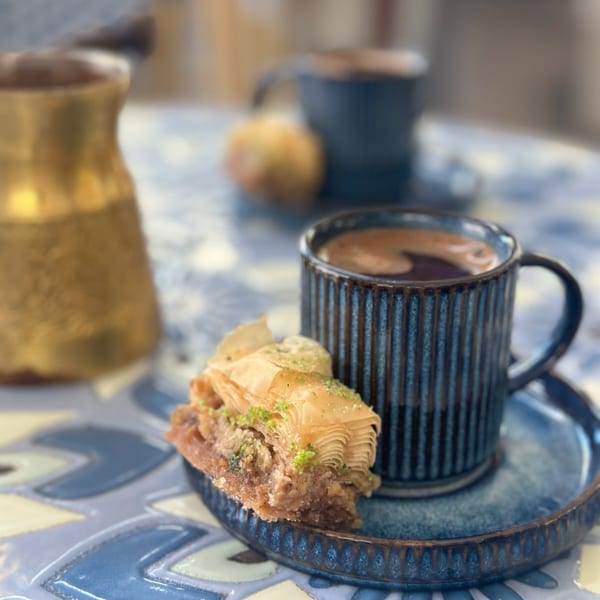 A slice of baklava with pistachios next to a dark blue cup of coffee on a patterned table.