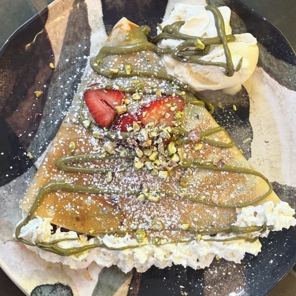 Manasiri Crepe with strawberries, pistachios, matcha drizzle, whipped cream, and powdered sugar on a dark plate.